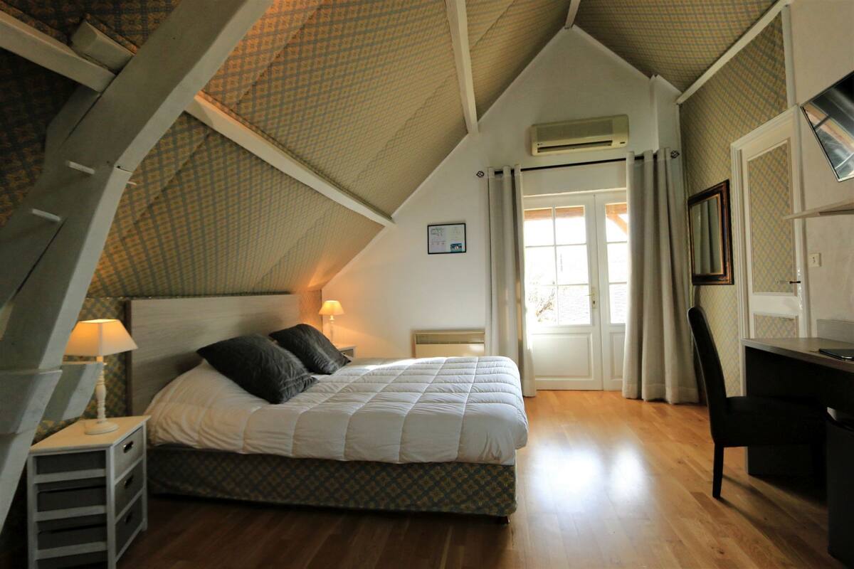 A bedroom with a bed, a desk, and a chair. There is a window with curtains and an air conditioning unit on the wall.