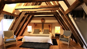 A cozy attic bedroom with a sloped ceiling, wooden beams, a large bed with patterned pillows, two chairs, and two bedside lamps. - Hôtel de la Ferme Lamy (Meyrals)