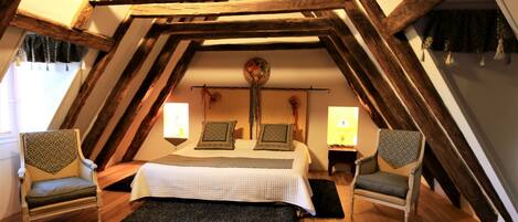 Un dormitorio acogedor en el ático, con techo inclinado, vigas de madera, una cama grande con almohadas estampadas, dos sillones y dos lámparas de noche.