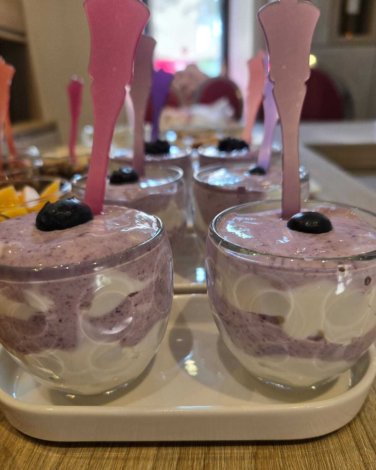Una bandeja con cuatro vasos que contienen postre en capas de color púrpura, coronado con una baya negra y un adorno rosa.