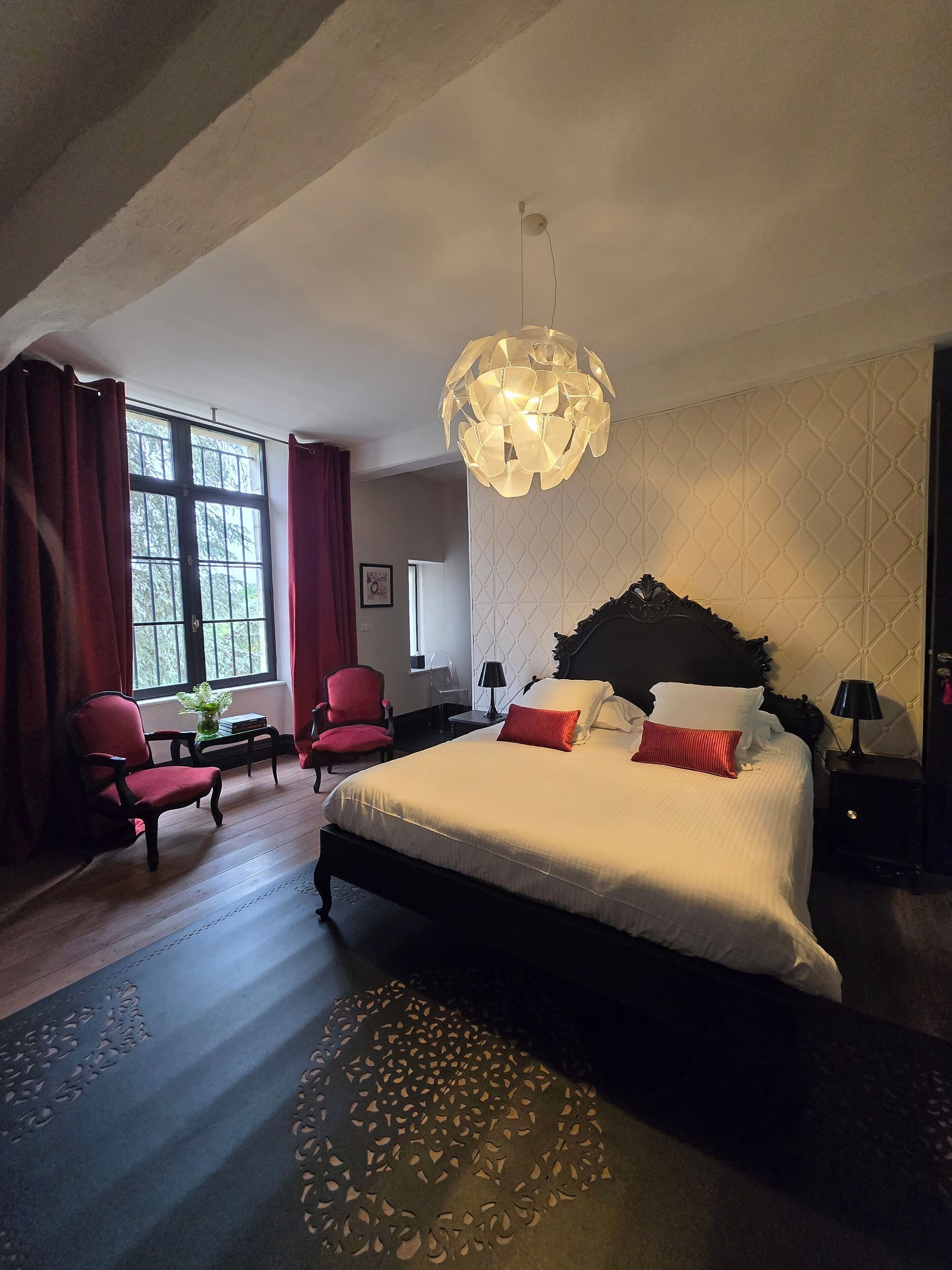 A hotel room with a large bed, two red chairs, a small table, a decorative lamp, and a unique ceiling light fixture.