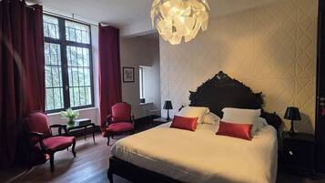 A hotel room with a large bed, two red chairs, a small table, a decorative lamp, and a unique ceiling light fixture.