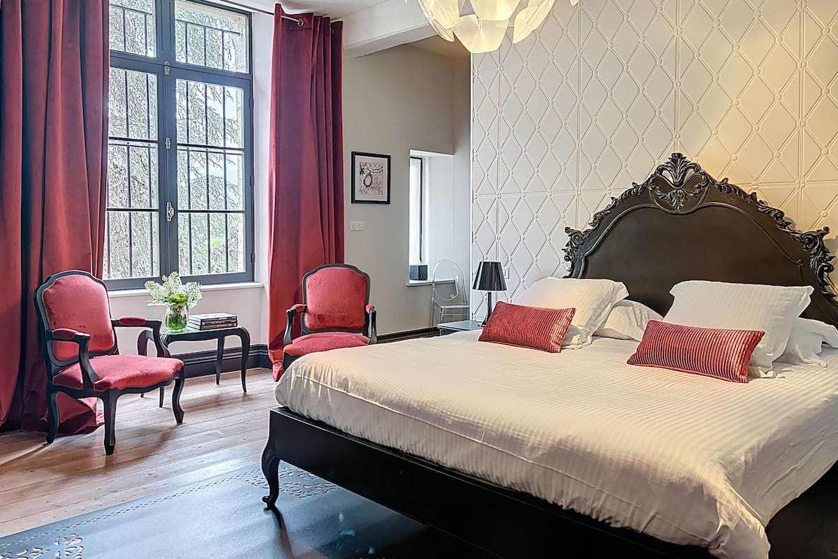 A hotel room with a large bed, red chairs, a small table with a vase, and a window with curtains.