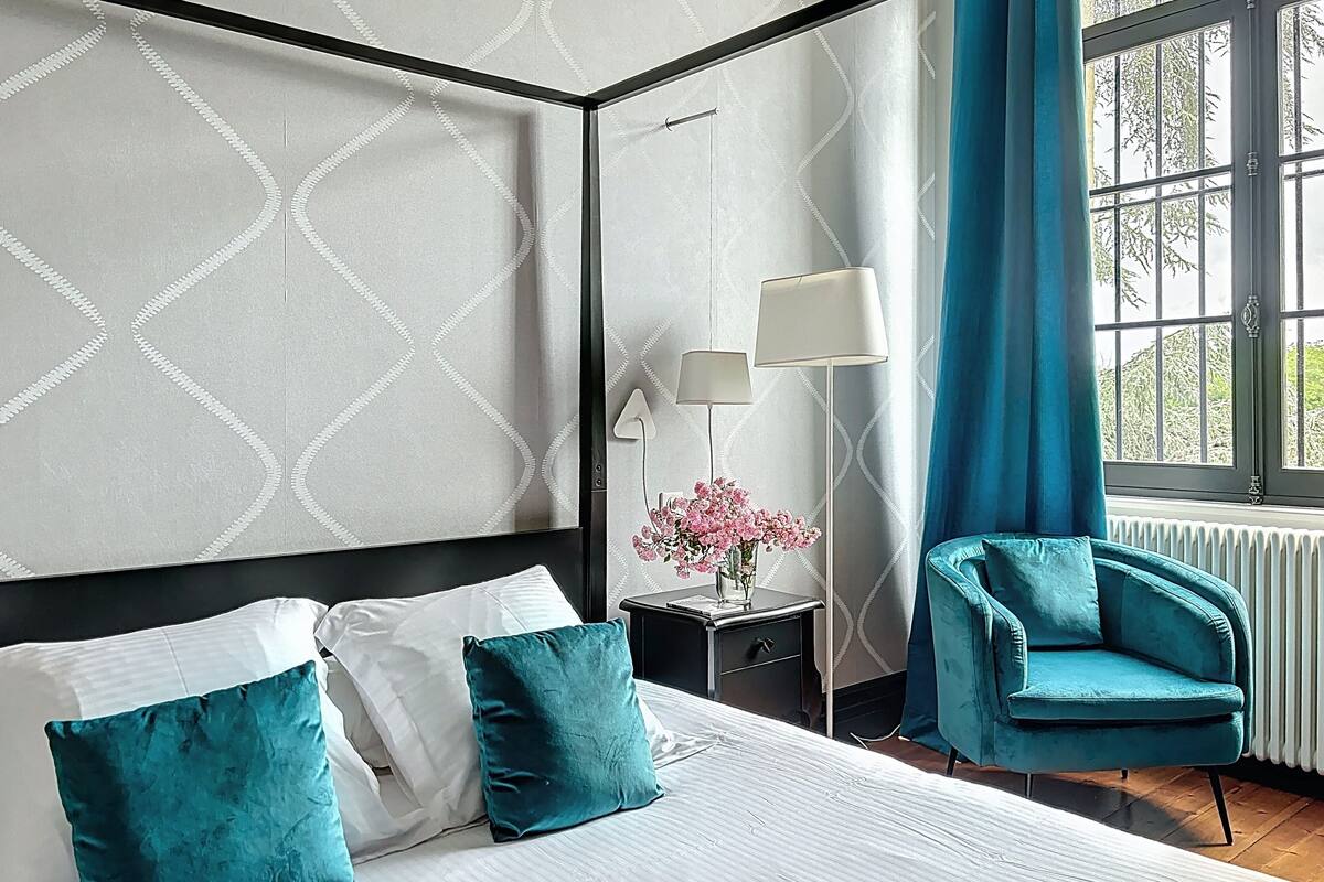 A modern hotel room with a large bed, a nightstand, a lamp, a chair, and a window with a view of trees.