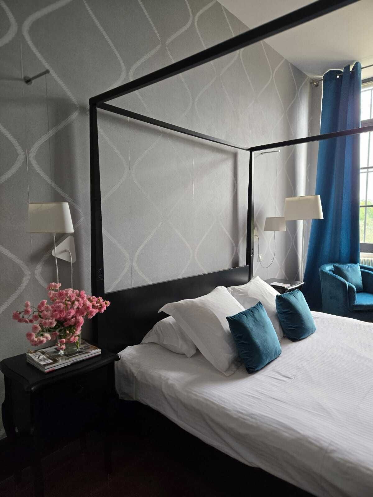 Un lit à baldaquin avec du linge de lit blanc et des oreillers bleus, une table de chevet avec une lampe et un vase rempli de fleurs roses.