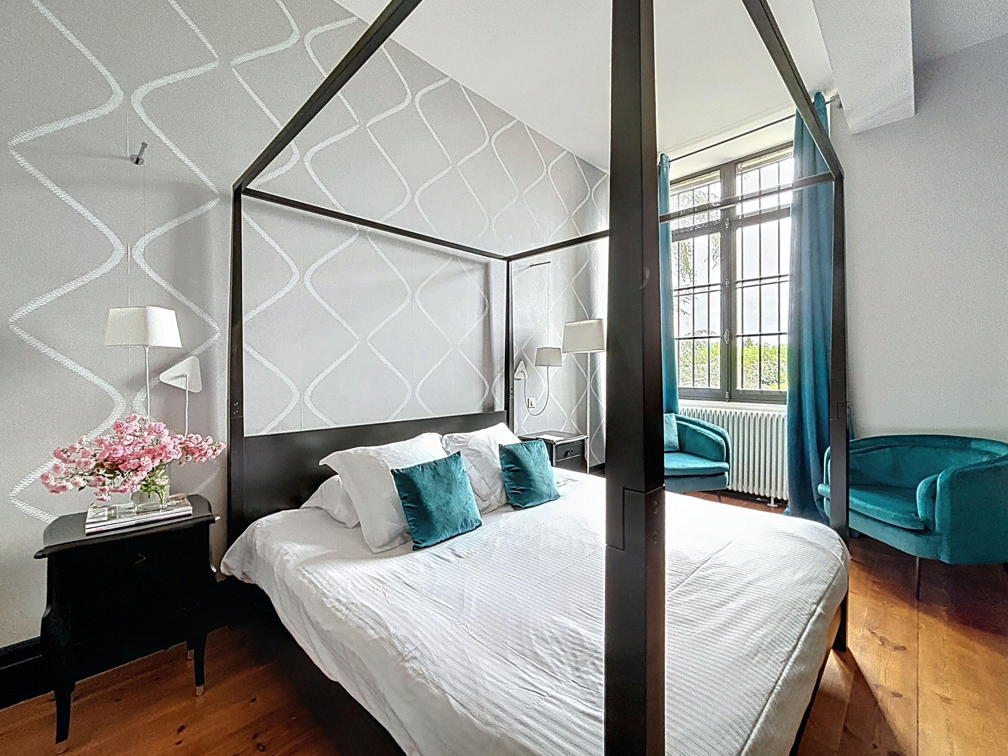 A modern bedroom with a four-poster bed, a nightstand with a lamp, a vase of flowers, and a view of the outdoors through a window.