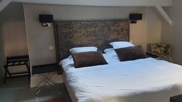 A neatly made bed with white linens and two patterned pillows, a small bedside table with a lamp, and a chair with a colorful backrest.