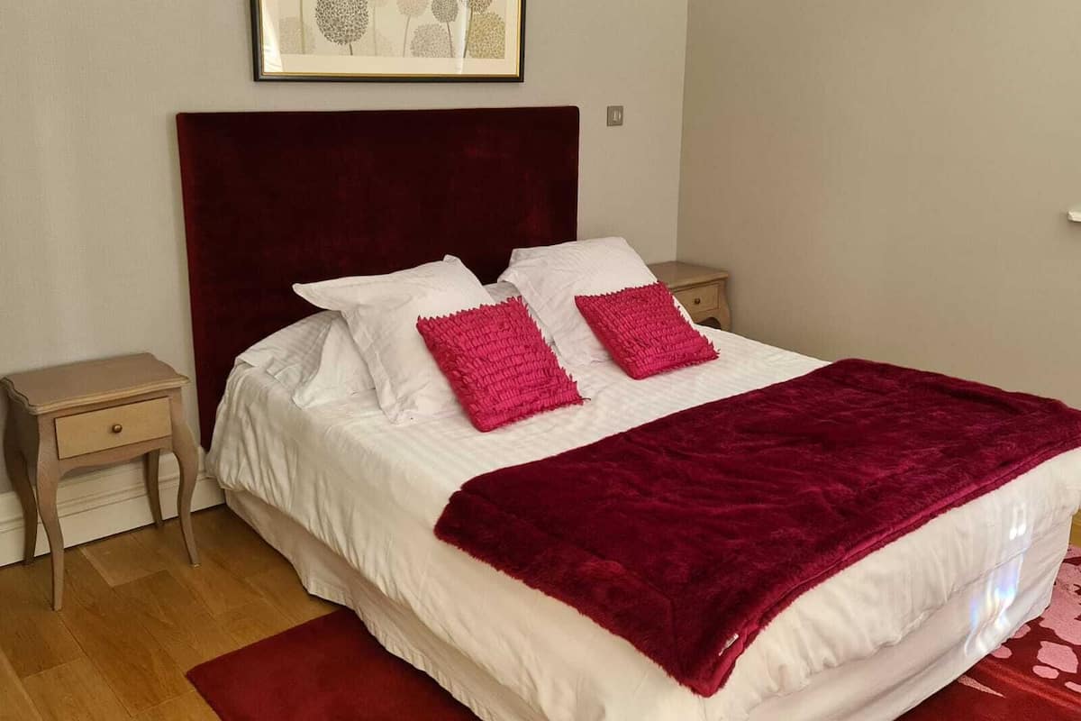 A neatly made bed with white linens and two red pillows, a dark red headboard, and a framed picture above.