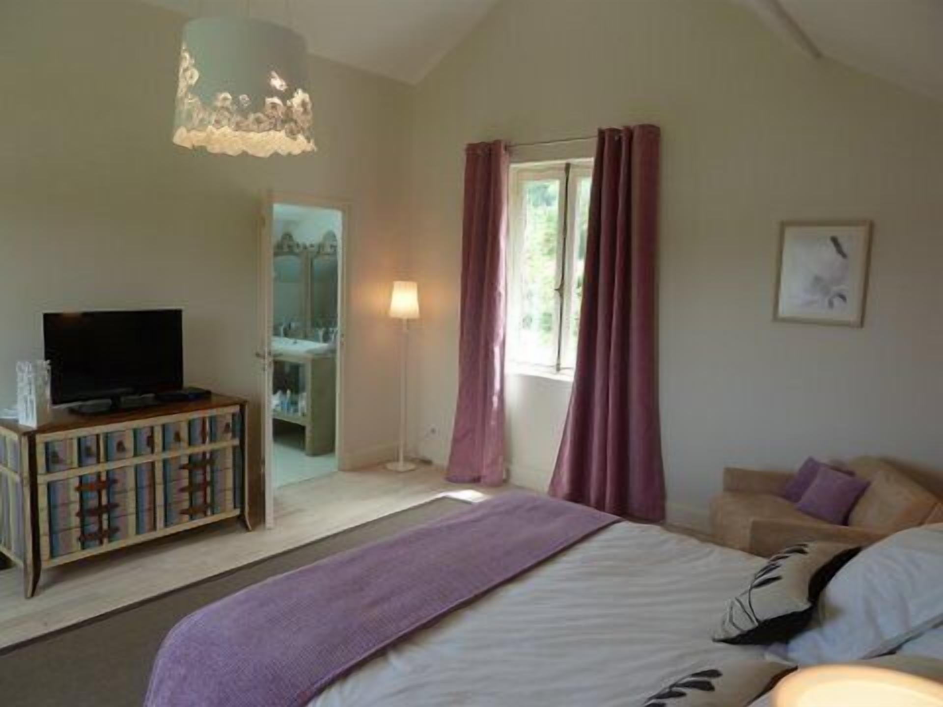 A bedroom with a bed, a television on a wooden cabinet, a floor lamp, and a window with curtains.