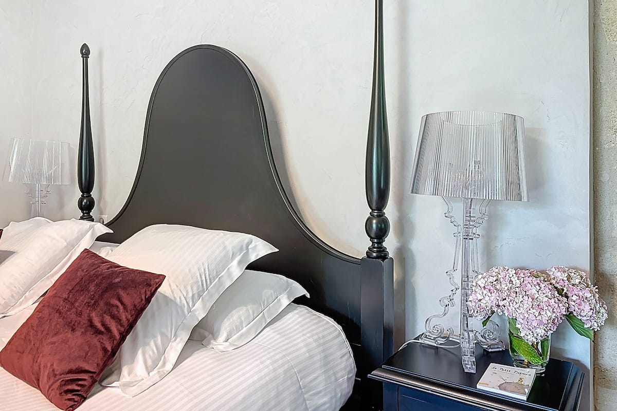 A neatly made bed with white and brown pillows, a dark headboard, a bedside table with a lamp, and a card.