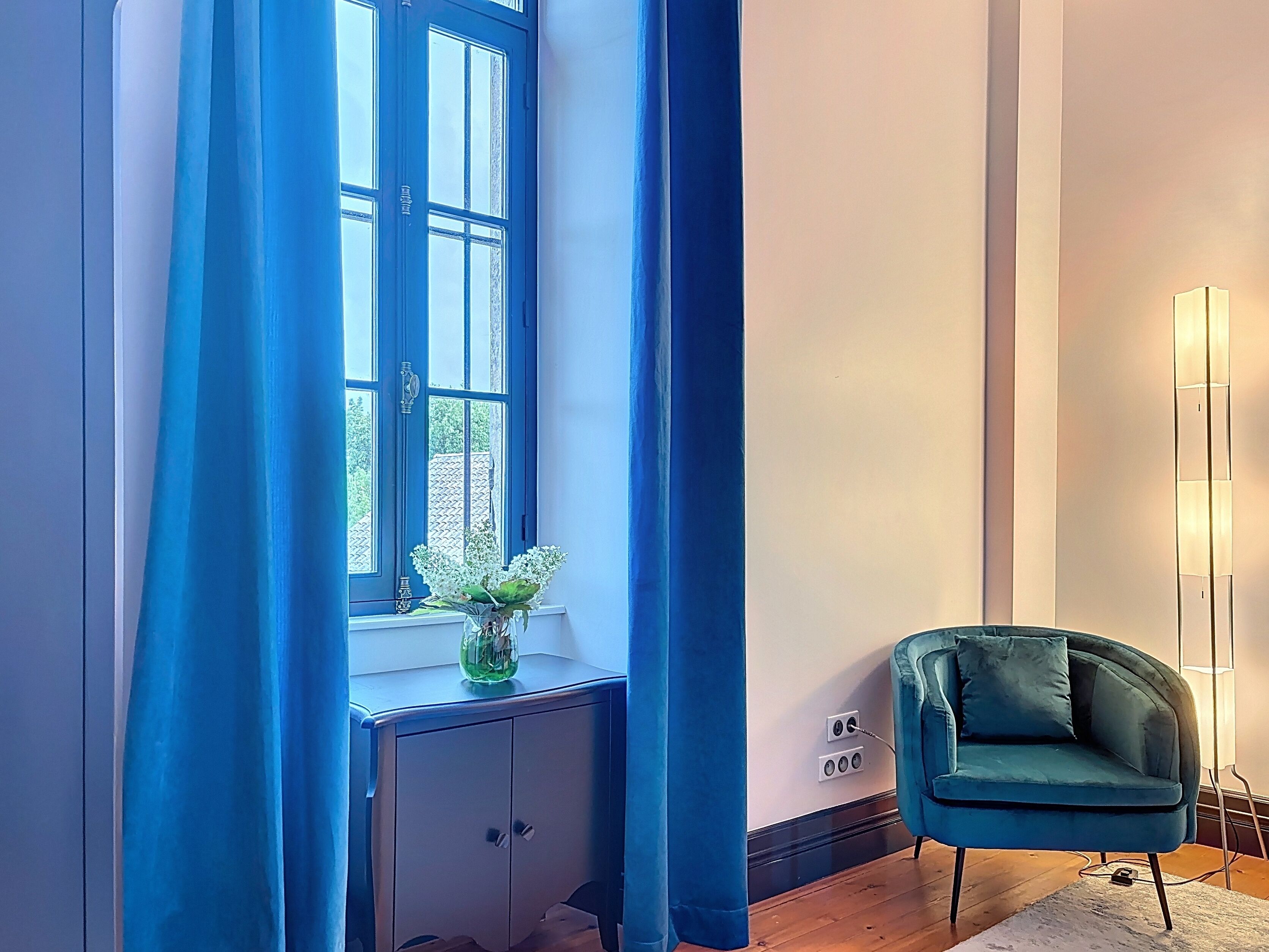 Une pièce avec une fenêtre, une chaise bleue et un vase rempli de fleurs, posé sur une commode.