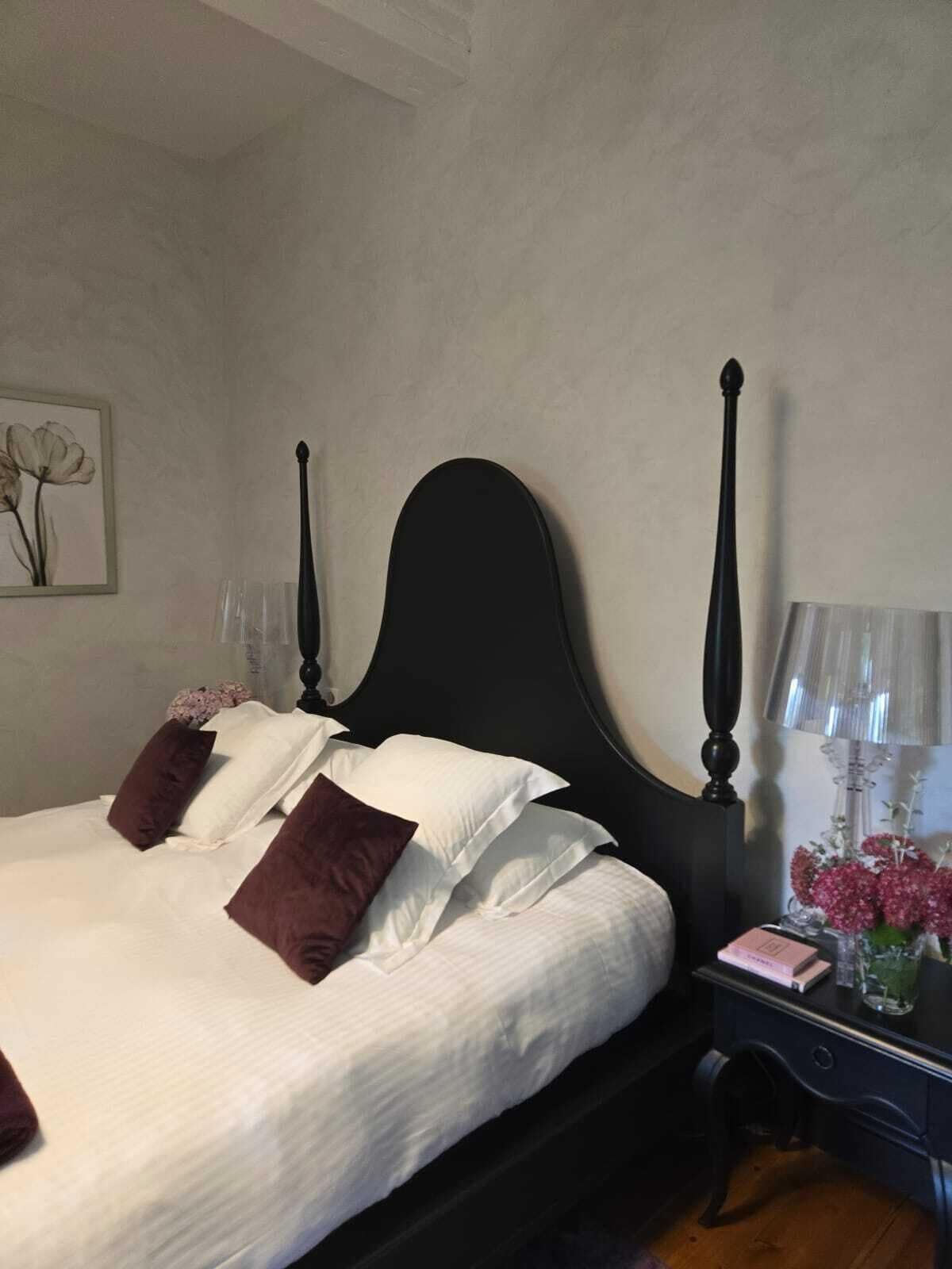 A neatly made bed with white and brown bedding, a dark headboard, and a bedside table with a lamp and a vase of flowers.