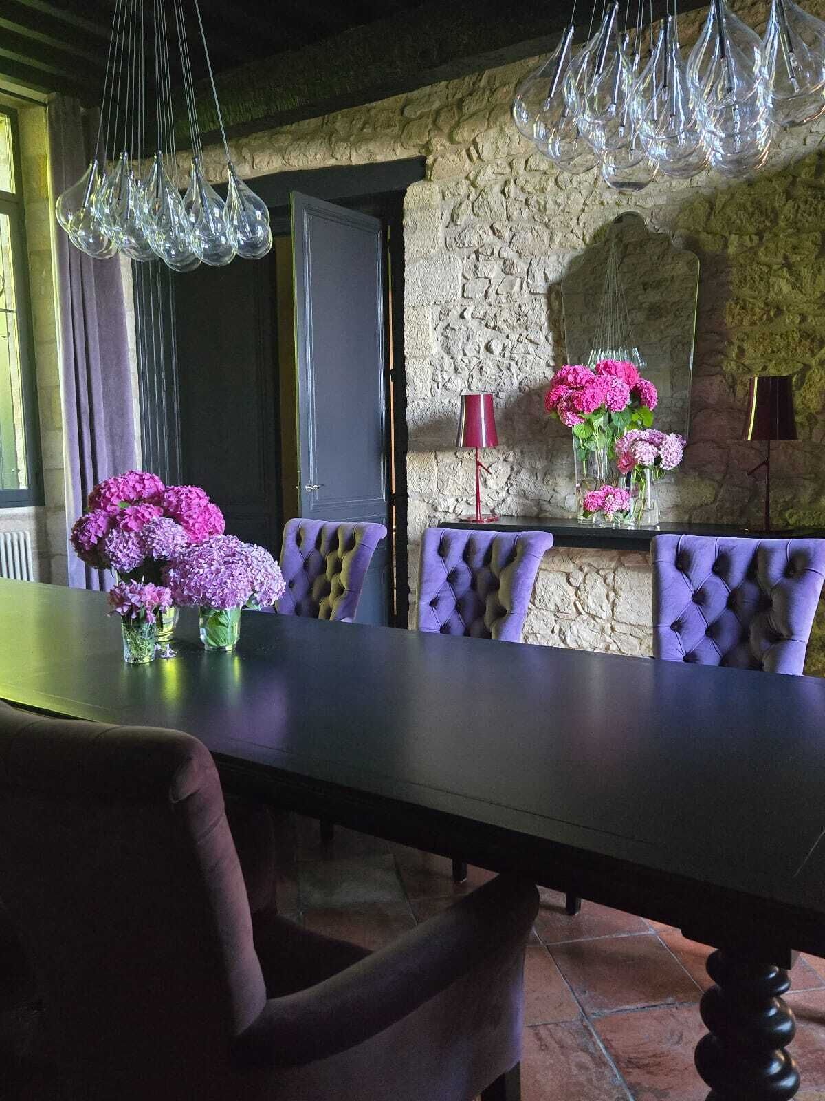 A dining area with a stone wall, a wooden table, purple upholstered chairs, and hanging glass light fixtures.