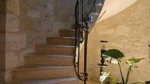 Una escalera de piedra con barandilla de hierro forjado, un pequeño ventanal arqueado y una planta en maceta.