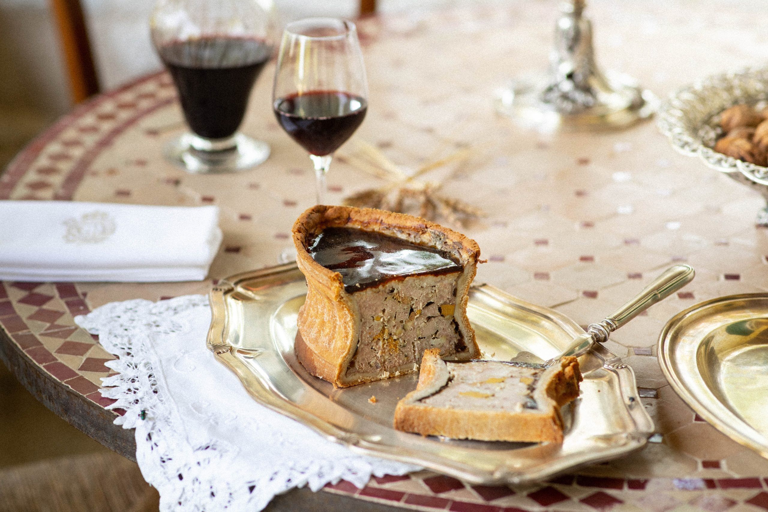 A slice of meat pie on a silver serving tray with a knife, a glass of red wine, and a white menu on a table with a patterned tablecloth.