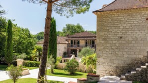 Un bâtiment en pierre avec un toit en tuiles, entouré d’un jardin bien entretenu où poussent différentes espèces de plantes et qui est agrémenté d’un sentier.