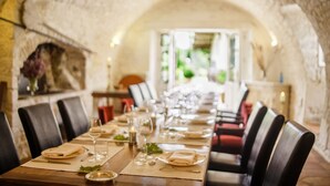 Une longue table à manger, dressée avec des assiettes, des verres et des couverts, est entourée de fauteuils en cuir, dans une pièce aux murs de pierre qui offre une vue sur la verdure environnante.