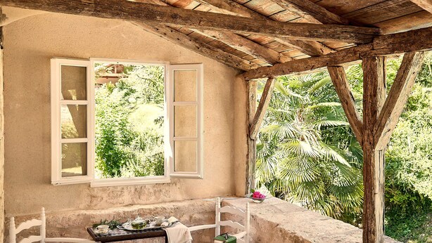 Un espace extérieur rustique aménagé avec une table et des chaises, une fenêtre aux volets ouverts et une vue sur une végétation luxuriante.