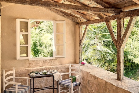 A rustic outdoor seating area with a table and chairs, a window with open shutters, and a view of lush greenery.