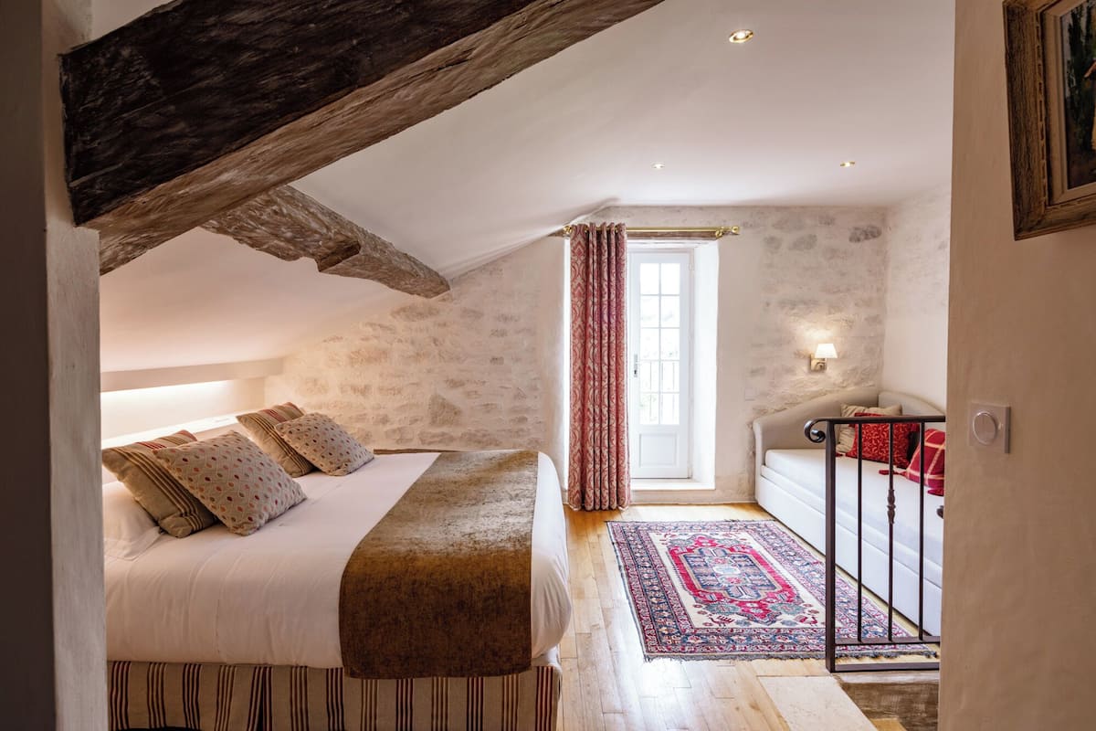 Een slaapkamer met een schuine kap, een bed met opvallende kussens, een rood-wit kleed en een klein balkon met een hekwerk.
