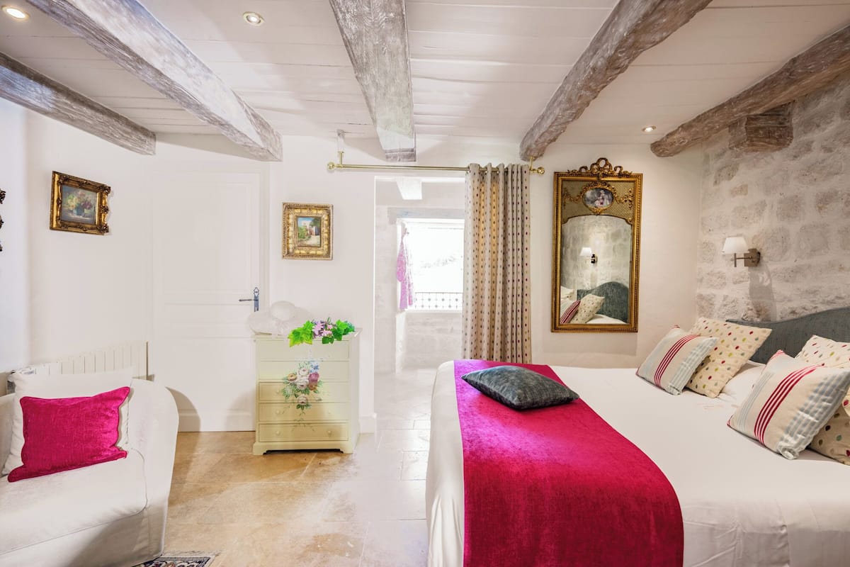Een slaapkamer met een bed, een dressoir en een spiegel. Er is een raam met gordijnen en een deur naar een andere kamer.