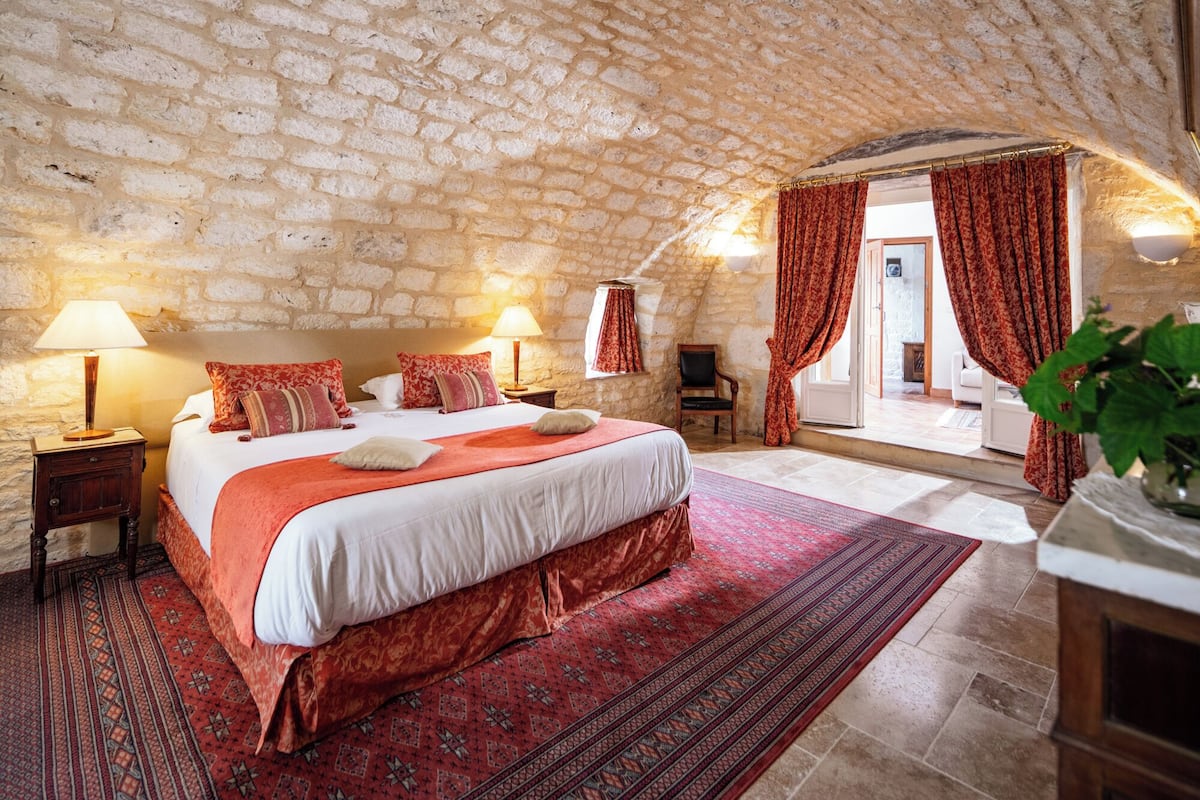 Een slaapkamer met een stenen plafond, een groot bed met rode beddengoed, twee nachtlampjes en een uitzicht op buiten door een deur.