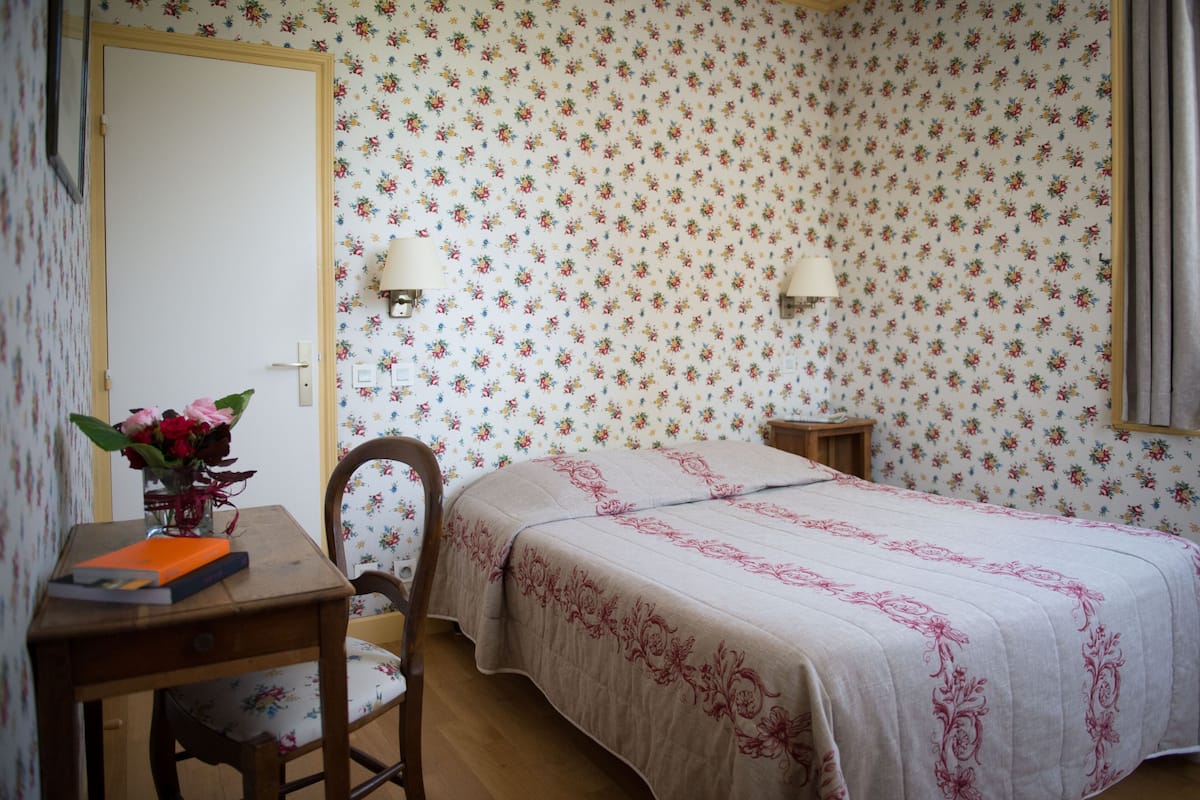 Un dormitorio con papel tapiz floral, una cama con colcha rosa, una mesita de noche de madera, una silla y un escritorio con un florero.