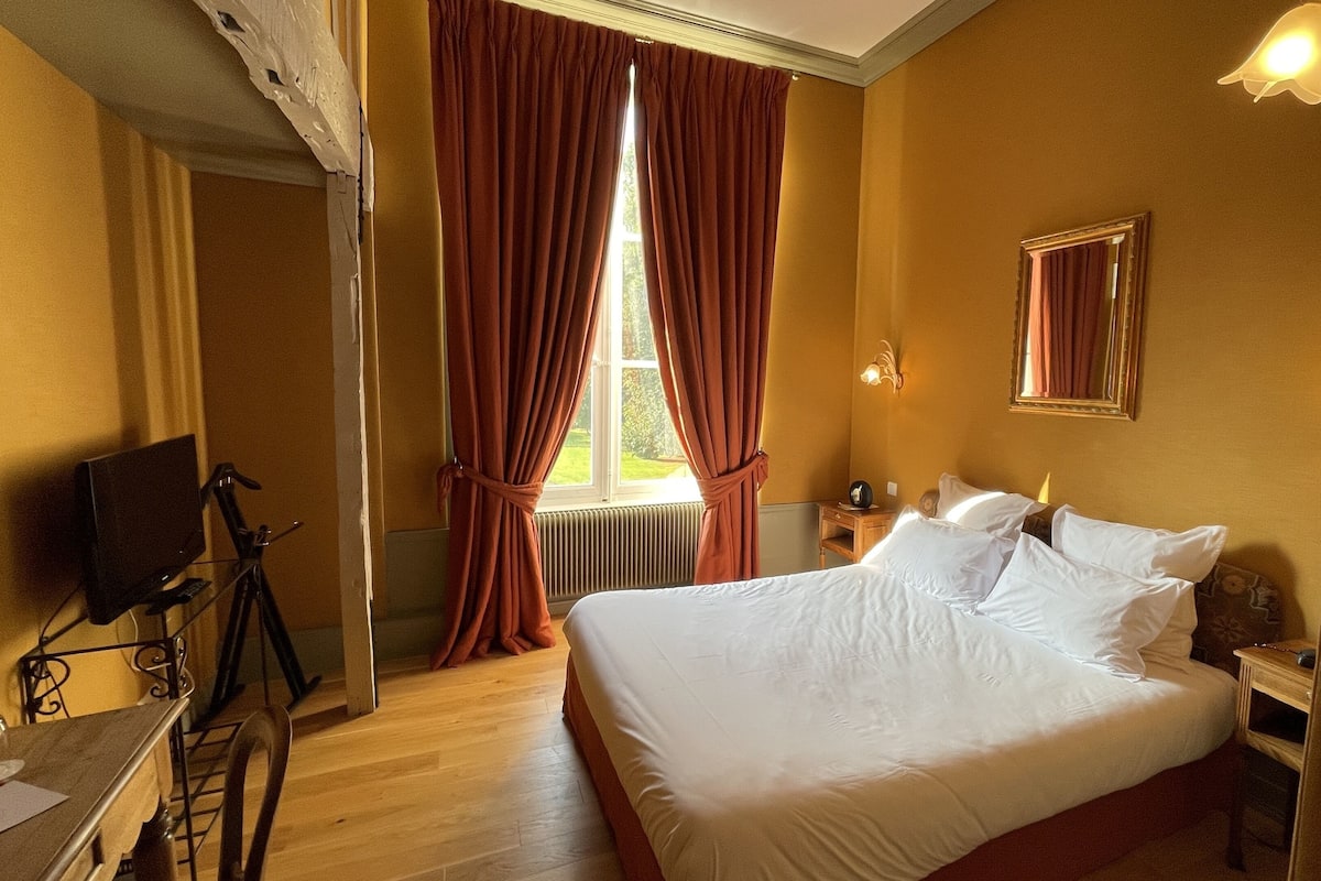 Una habitación de hotel con una cama grande, un televisor sobre un mueble, una ventana con cortinas rojas y un espejo en la pared.