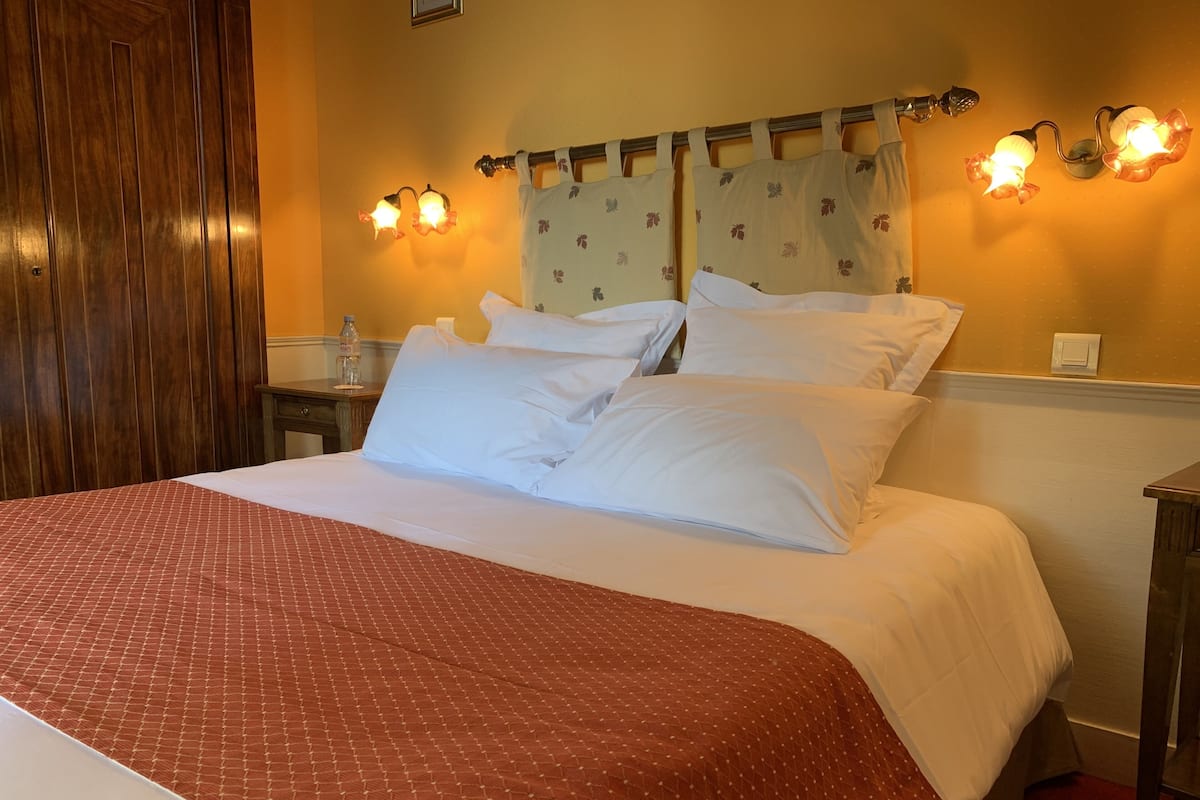 Habitación de hotel con una cama grande, ropa de cama blanca, cabecero de madera, una mesita de noche con una botella y luces fijadas a la pared.
