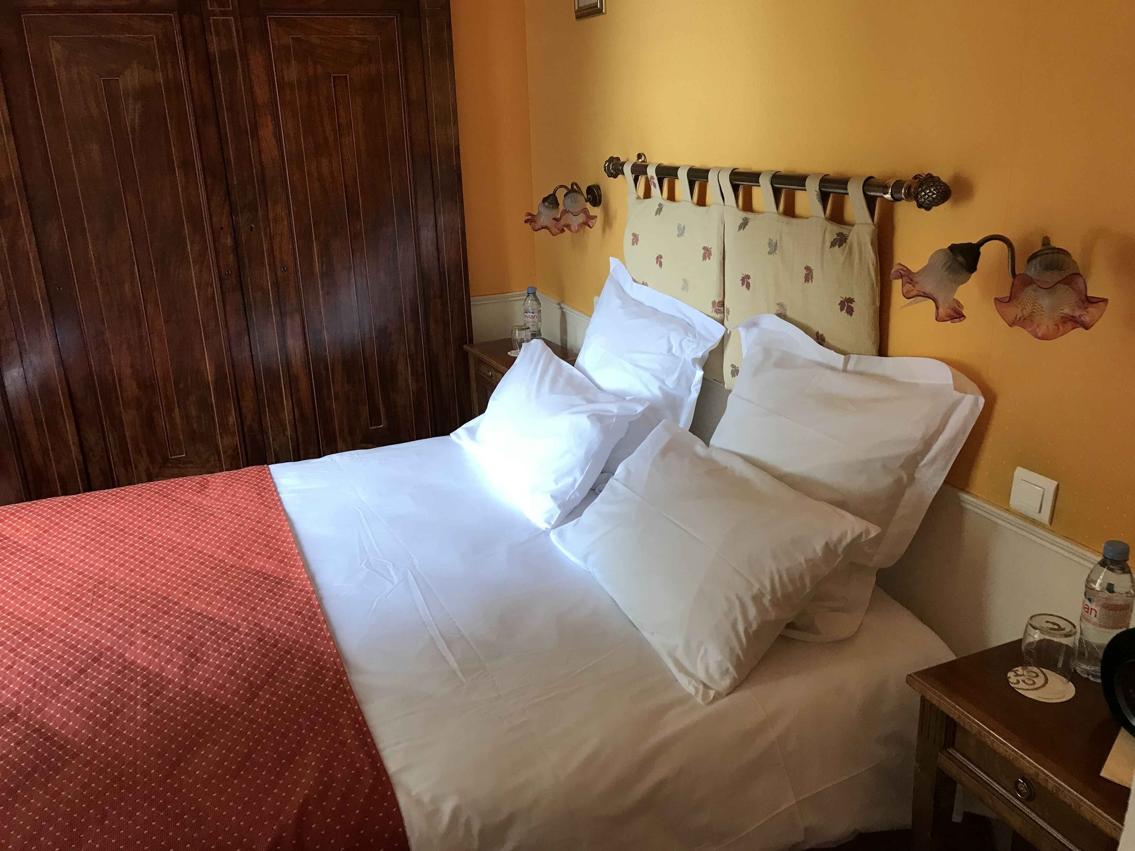 A neatly made bed with white linens, a wooden nightstand with a bottle and glasses, and a wall with a floral headboard.