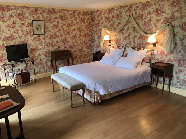 Une chambre à coucher comprenant un lit, des tables de chevet, un banc, un meuble pour la télévision et un mur recouvert d’un papier peint à motifs floraux.
