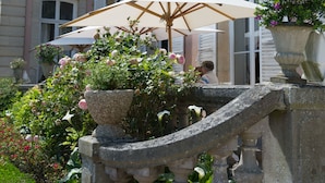 Een stenen balustrade met een witte parasol, een rozenstruik en een bloempot met paarse bloemen.
