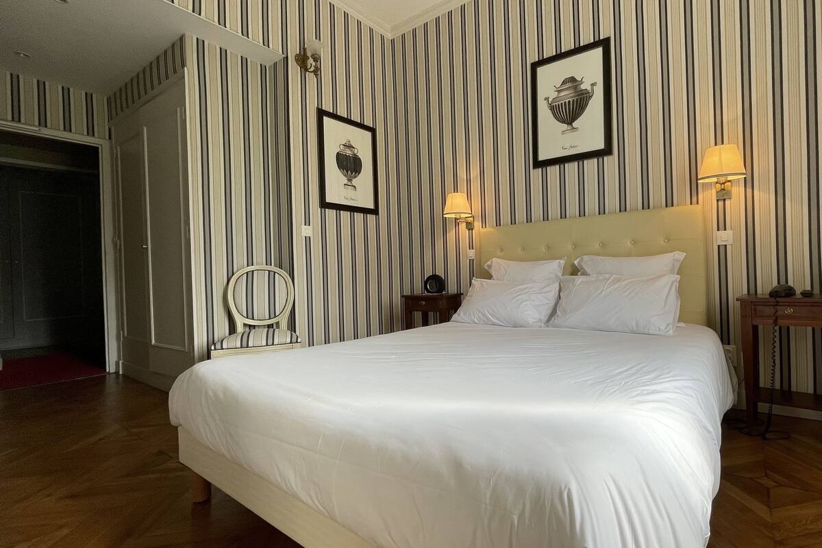 Habitación de hotel con una cama grande, papel tapiz a rayas, dos cuadros enmarcados, una mesita de noche con lámpara y una silla.