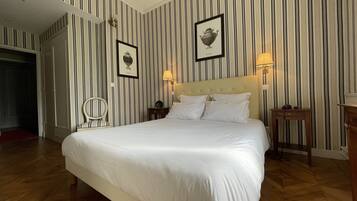 A hotel room with a large bed, striped wallpaper, two framed pictures, a nightstand with a lamp, and a chair.