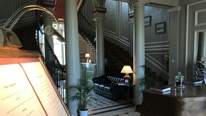 Een statige lobby met een zwart-wit schachbordvloer, een trap, een leren bank en een piano.