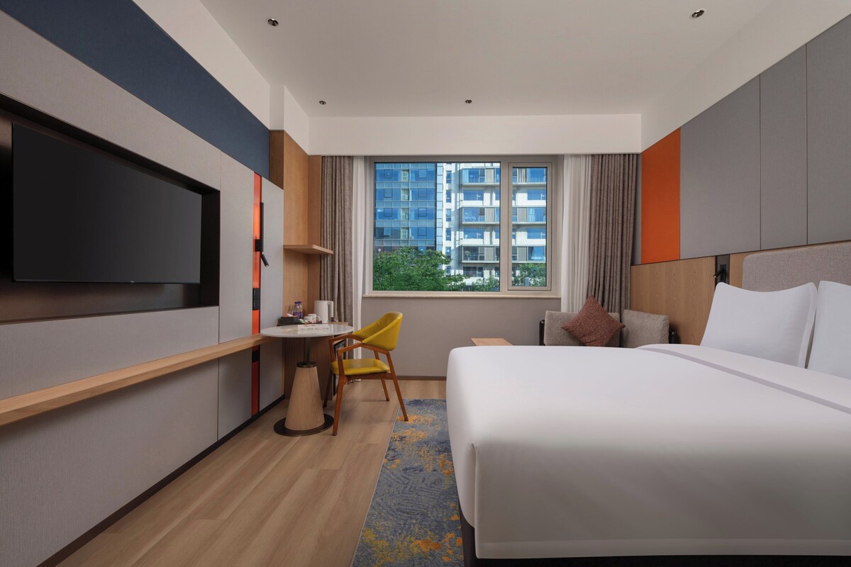 A modern hotel room with a large bed, a desk with a chair, a flat-screen TV, and a window with a city view.
