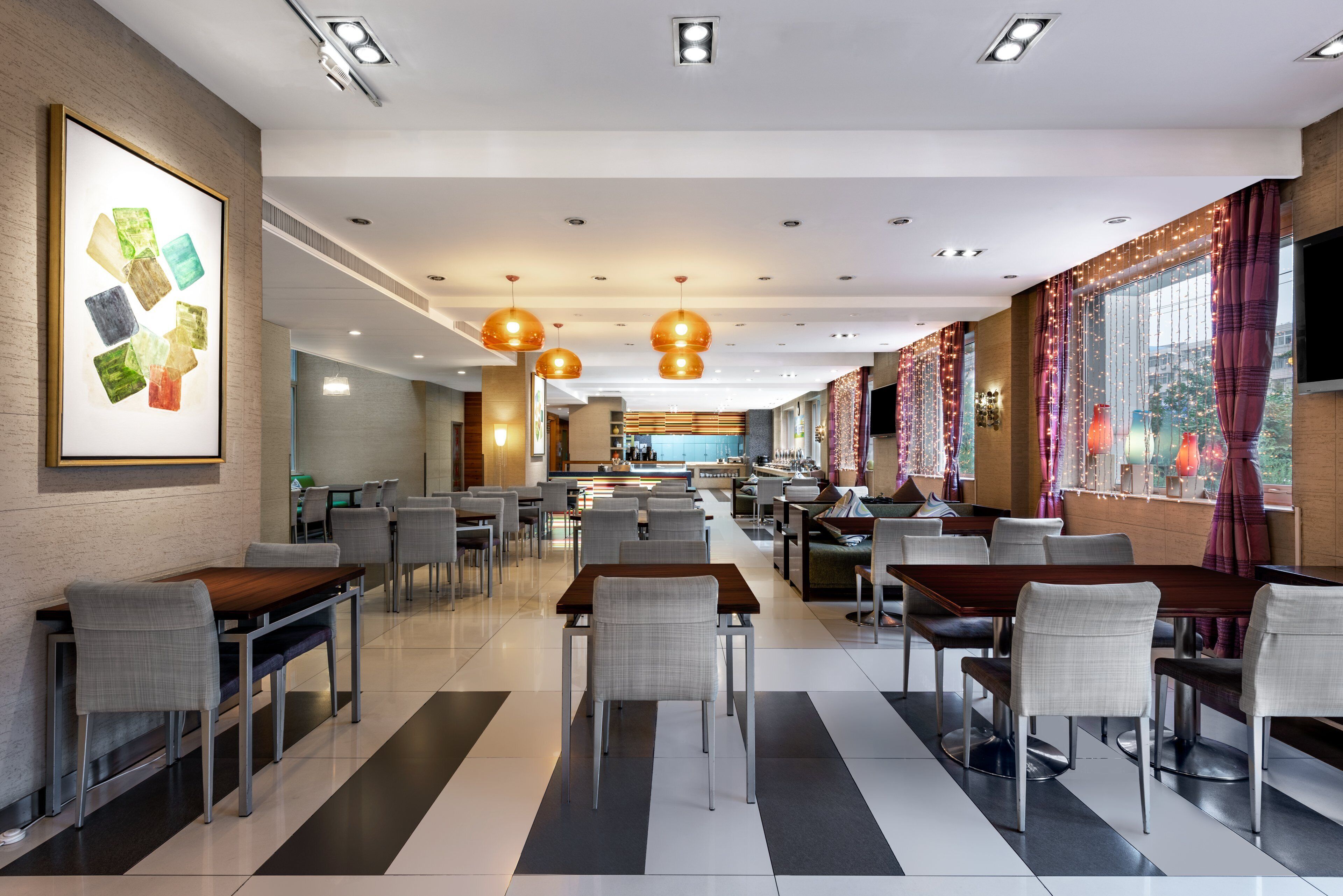 A modern restaurant with a long counter, multiple tables, and chairs. There is a large painting on the wall and a kitchen area in the background.