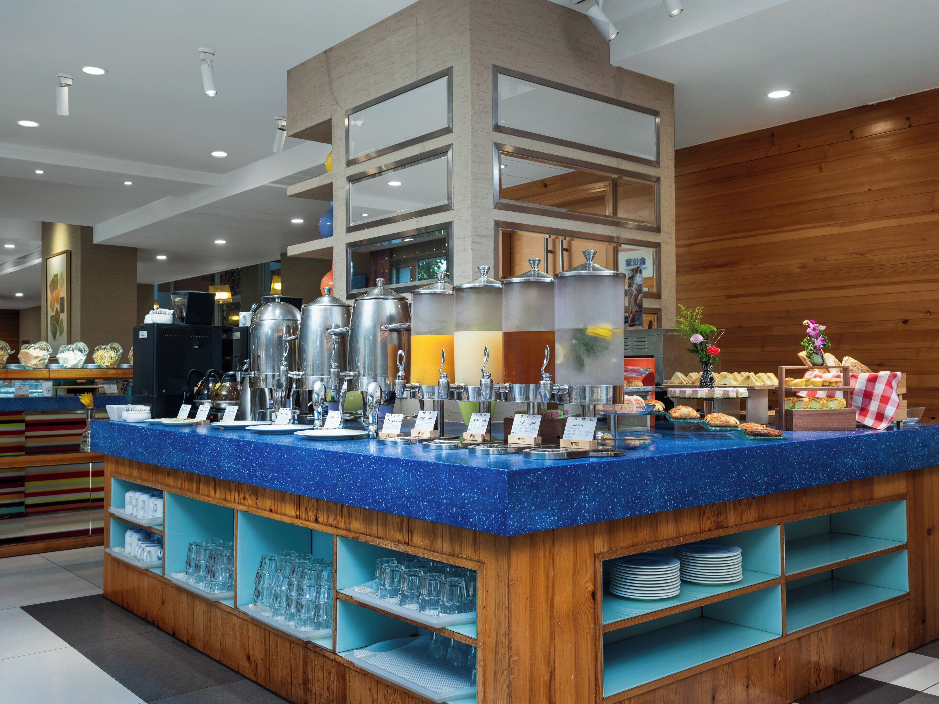 Un bar de petit-déjeuner doté de distributeurs en acier inoxydable, d’un comptoir bleu et d’étagères en bois où sont disposés des verres.