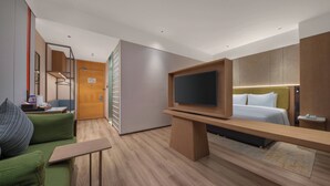 Een moderne hotelkamer met een houten vloer, een bed met wit beddengoed, een flatscreen televisie op een houten standaard, een groene bank met een kussen, en een houten garderobe.
