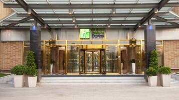 A hotel entrance with a glass facade, a green and blue sign, and potted plants.