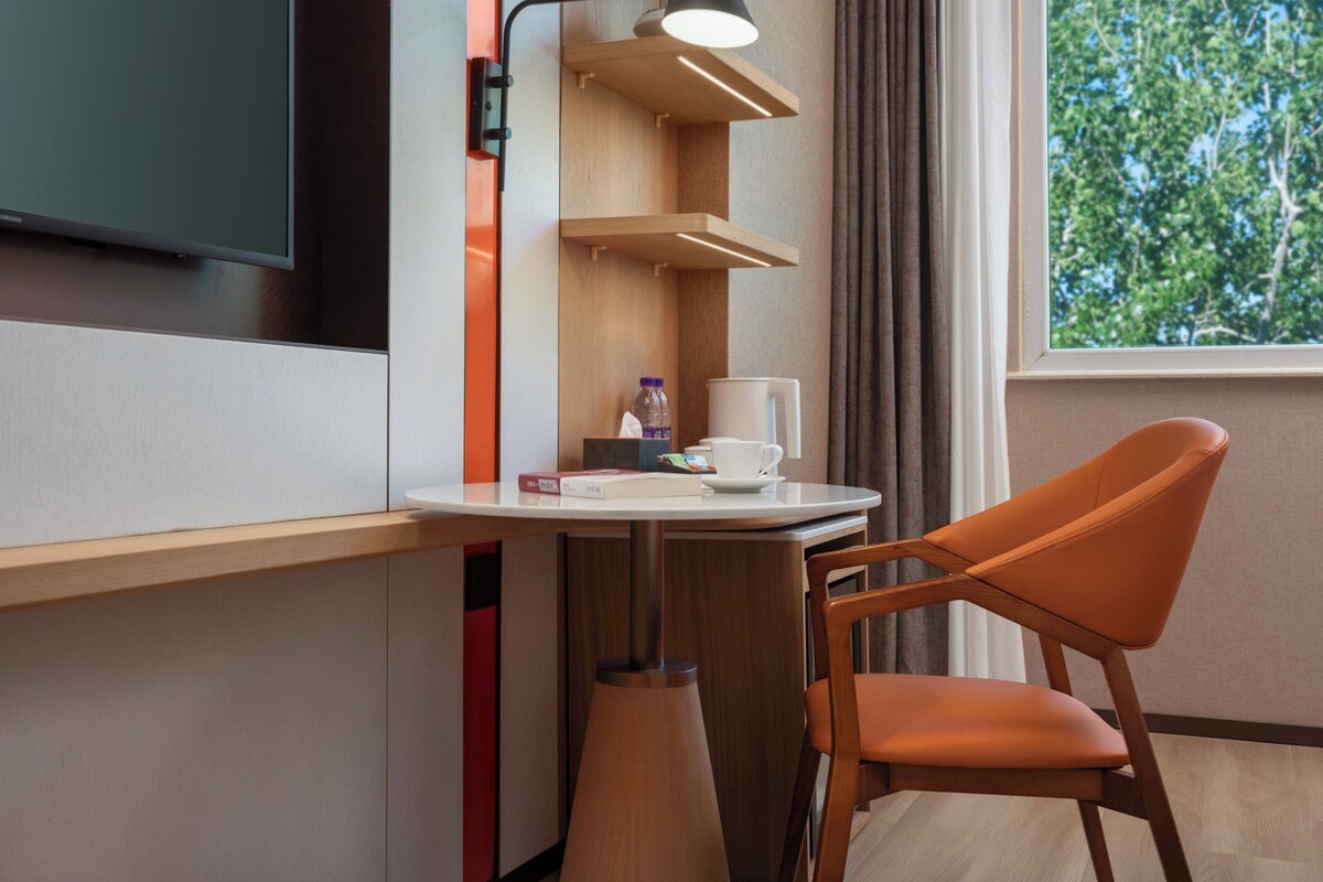 A modern hotel room with a flat-screen TV, a small table with a chair, a lamp, and a window with a view of trees.