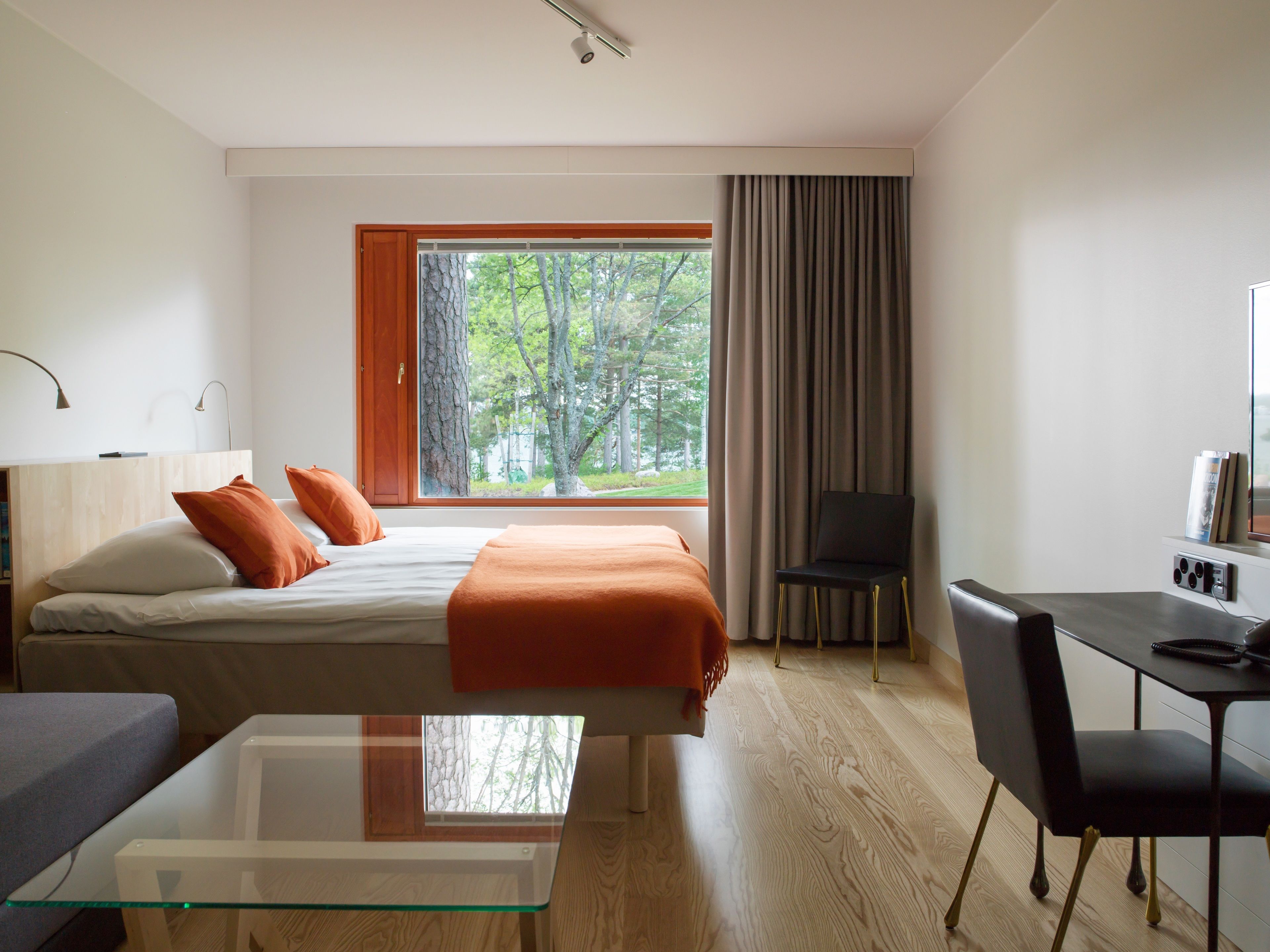 A modern hotel room with a large window, a bed with orange bedding, a desk with a chair, and a small table.
