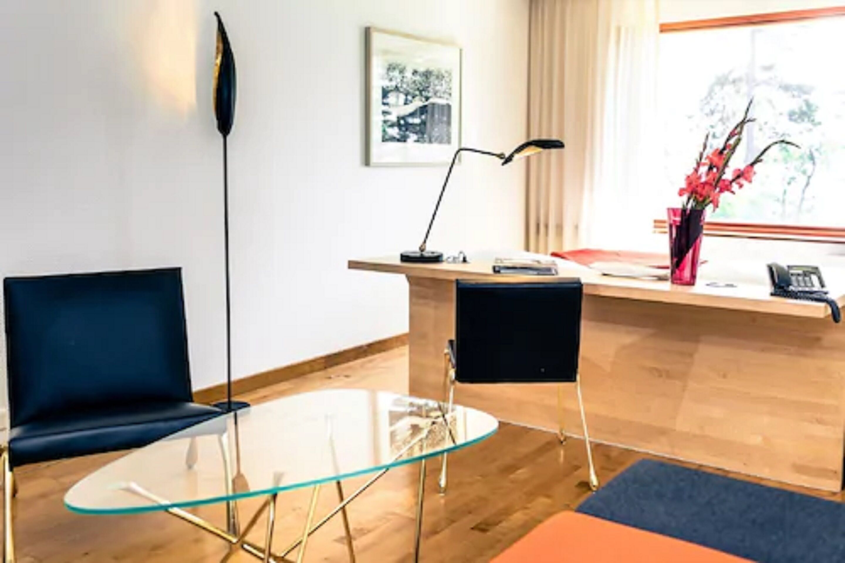 Moderni huone, jossa on lasipöytä, kaksi mustaa tuolia, puinen työpöytä, musta lattiavalaisin ja ikkuna, josta avautuu näkymä puihin.