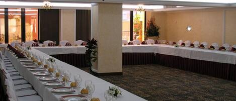Un salón de banquetes con largas mesas dispuestas para un evento formal, con manteles blancos, platos ordenados y cristalería.