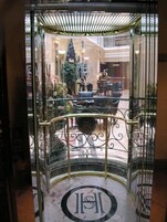Entrada de elevador de vidro com planta baixa circular, um elemento decorativo central e uma área de estar com cadeiras e uma mesa.