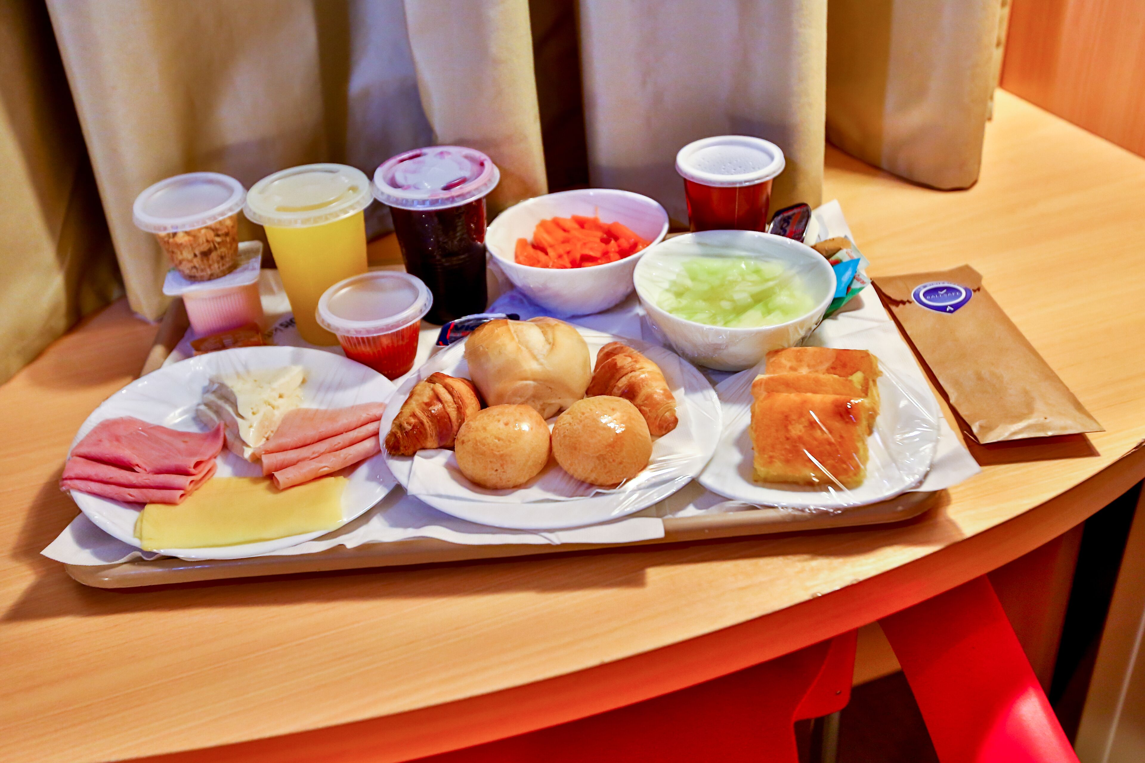 Una bandeja con diversos alimentos, incluyendo croissants, queso, jamón y verduras, junto con varias bebidas.