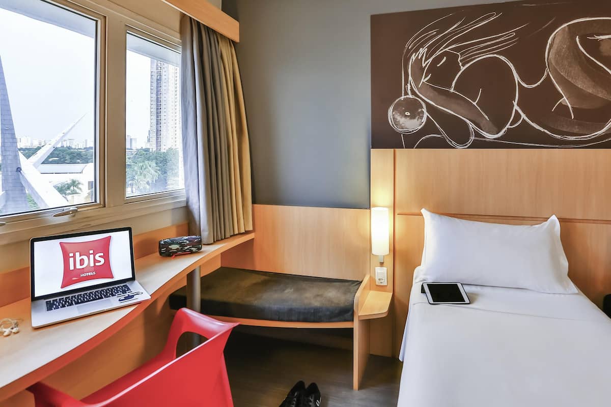 Ein Hotelzimmer mit Bett, Schreibtisch mit Laptop, Stuhl, Nachttisch mit Lampe und Blick auf Gebäude.