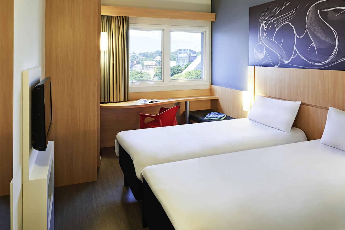 Ein Hotelzimmer mit zwei Betten, einem Schreibtisch, einem Stuhl, einem Fernseher und einem Fenster mit Blick auf Gebäude.