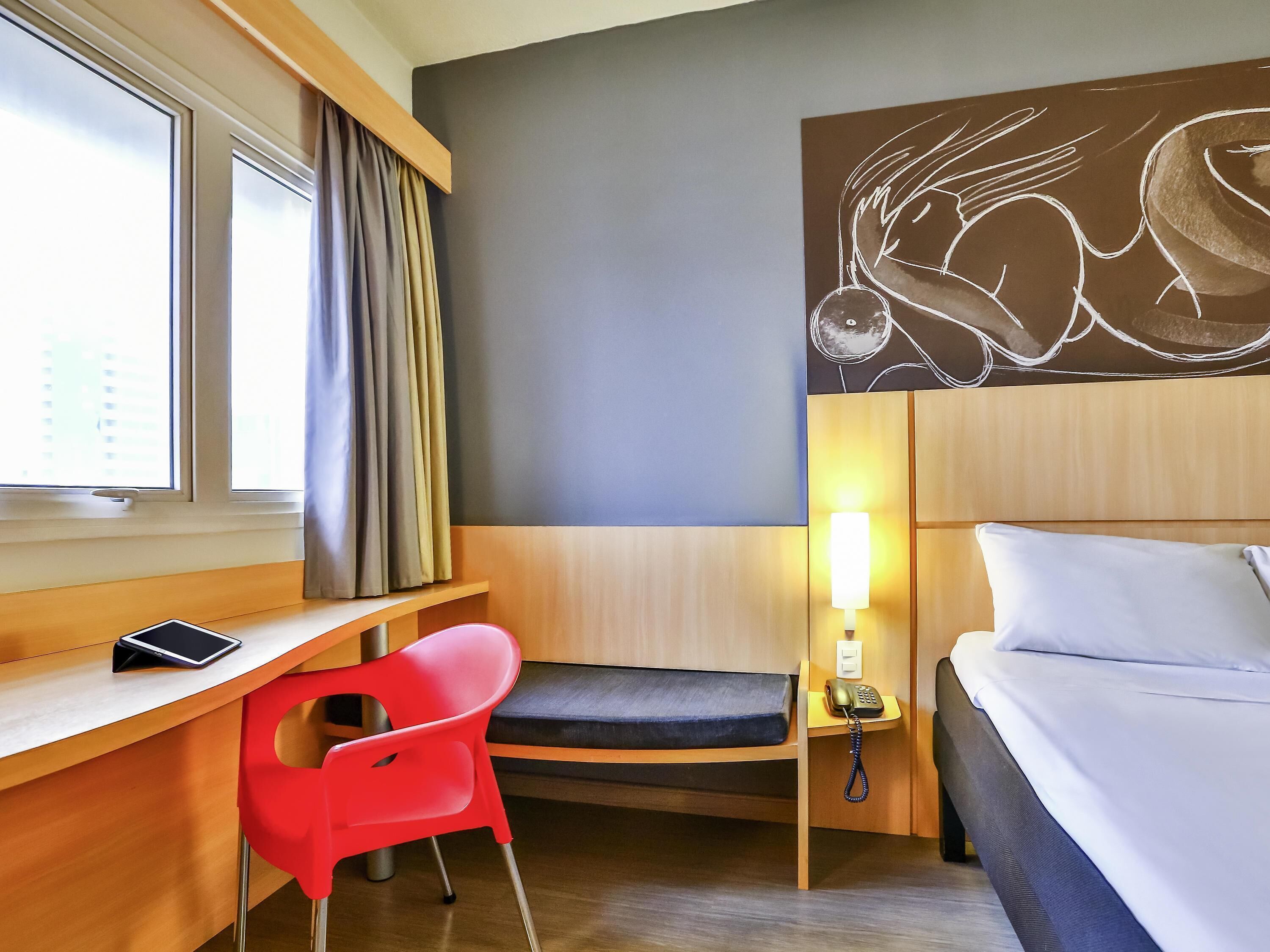 Quarto de hotel com cama, escrivaninha com cadeira, mesa de cabeceira com abajur e uma obra de arte na parede.