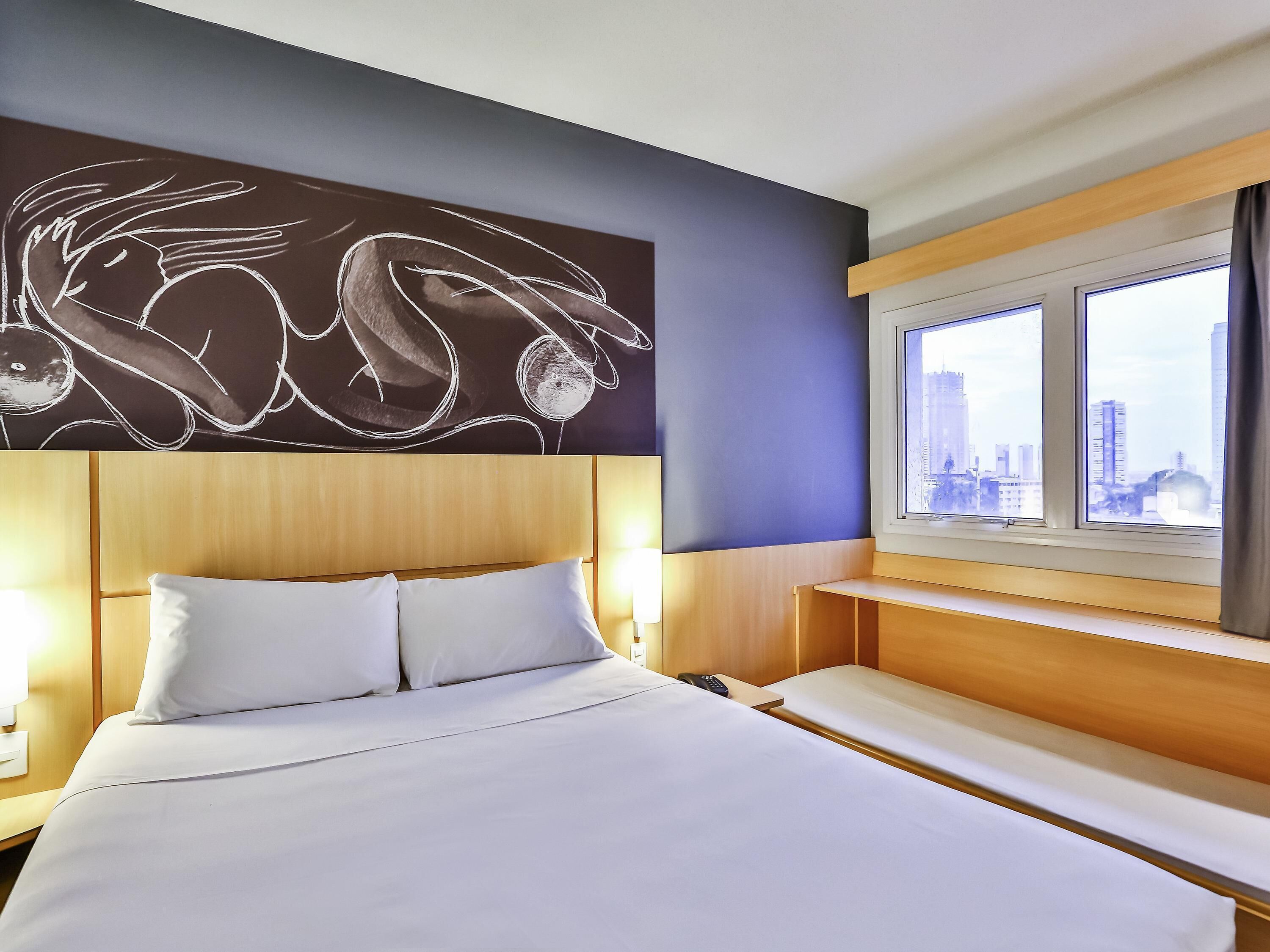 Habitación de hotel con cama, mesitas de noche, un amplio ventanal y una obra de arte en la pared.